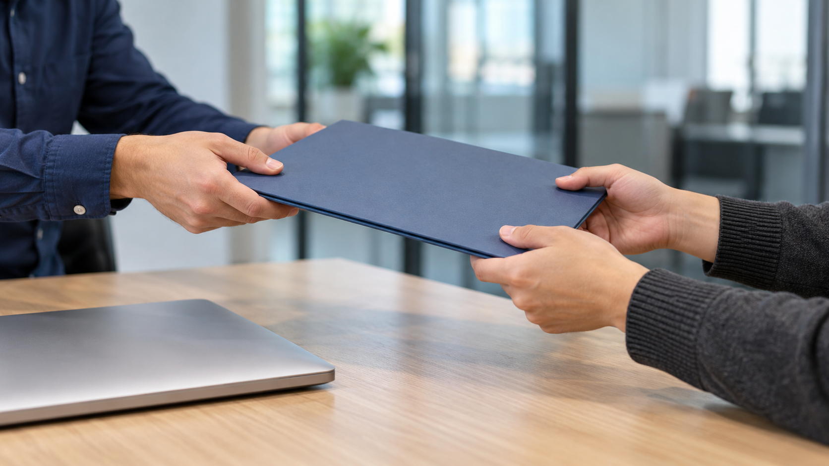 Colleagues handing over a plain document folder beside a closed laptop