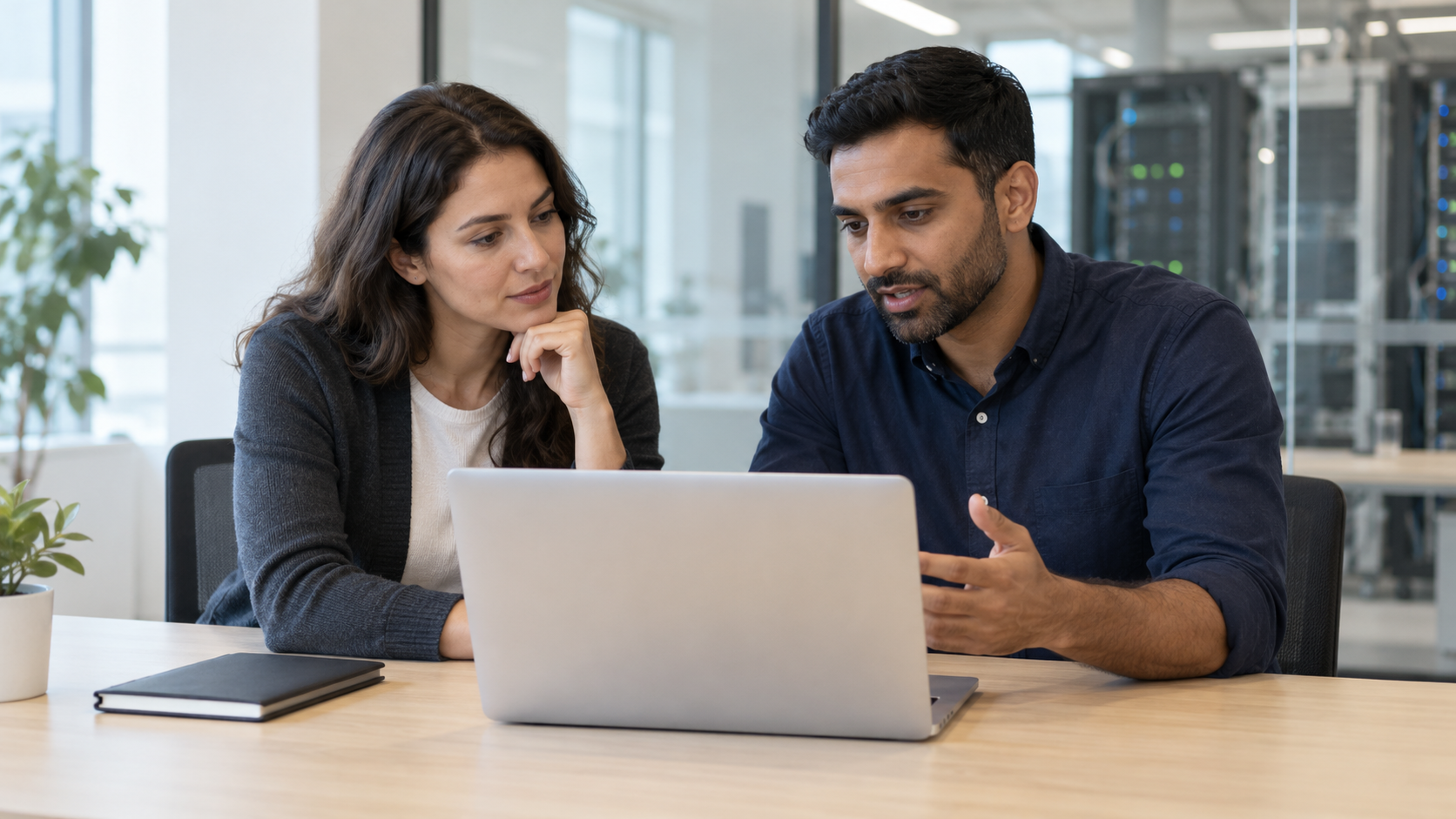 Two colleagues discussing maintenance decisions beside a laptop in an office near an equipment room